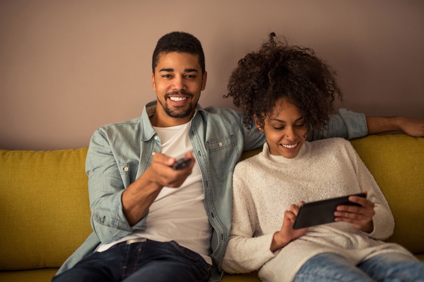 Junges lächelndes Paar auf dem Sofa vor Tablet und Fernseher